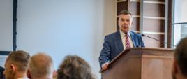 Minister Wieczorek stoi za mównicą i mówi do dwóch mikrofonów, przed nim sala wypełniona siedzącymi na krzesłach ludźmi.