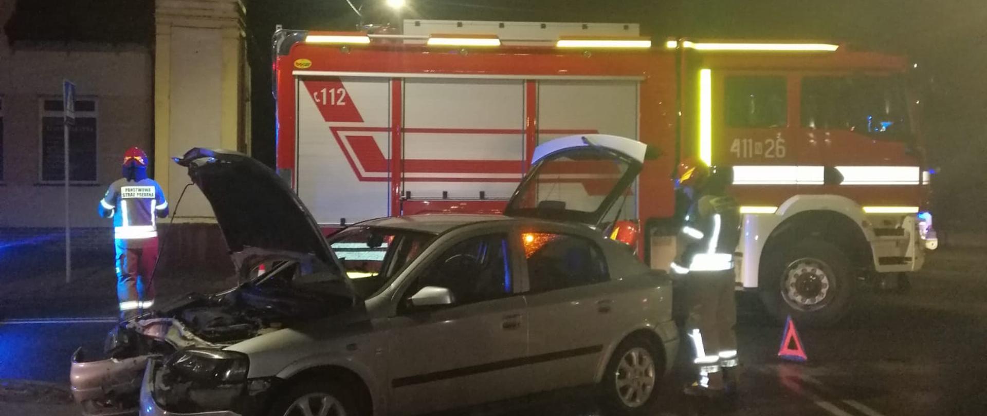Zdjęcie zdobione nocą. Uszkodzone srebrne auto z otwarta maska i klapą tylną. Za pojazdem stoi samochód pożarniczy i strażacy.