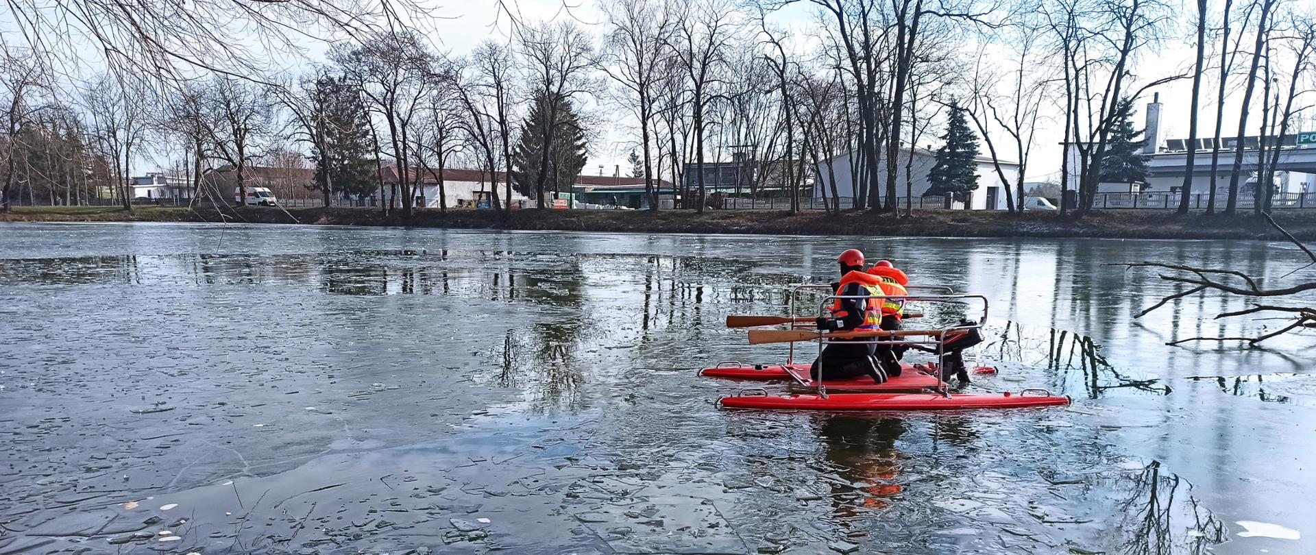 Ćwiczenia z zakresu ratownictwa na lodzie 2023 - sokołowscy strażacy doskonalili się z zakresu ratowania osób tonących po załamaniu tafli lodowej na stawie, 2 strażaków na czerwonym katamaranie pływa po stawie.