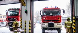 W dwóch bramach garażowych stoją dwa czerwony strażackie wozy bojowe. Przed każdym stoi strażak w żółtym mundurze bojowym.