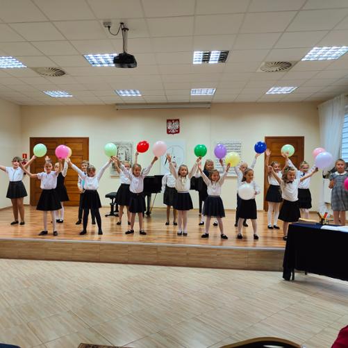 Zdjęcie przedstawia scenę sali koncertowej PSM w Grajewie, na której stoją uczniowie klas pierwszych ubrani na galowo. Dzieci trzymają nad głowami kolorowe baloniki. 