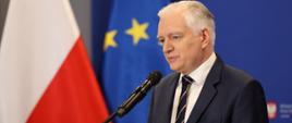 Wicepremier i minister rozwoju, pracy i technologii Jarosław Gowin stojący przy mównicy na konferencji prasowej, po jego lewej stronie flagi Polski I UE