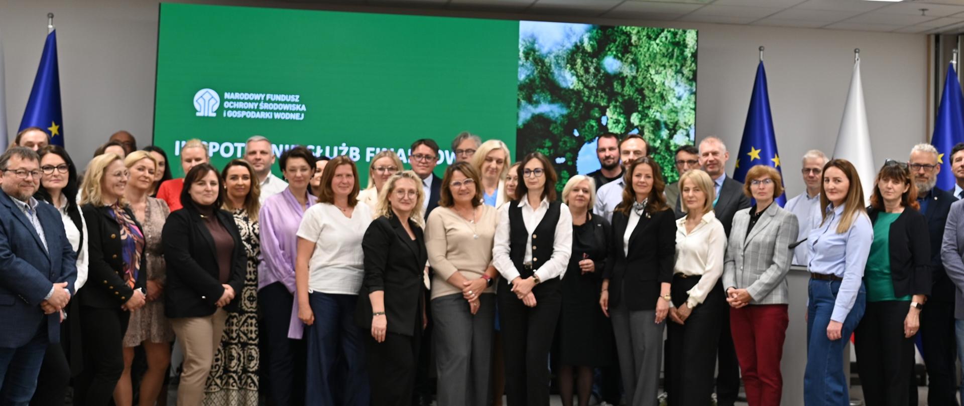 Grupowe zdjęcie uczestników III Spotkania Służb Finansowych NFOŚiGW i WFOŚiGW. Uczestnicy stoją w sali konferencyjnej przed ekranem z logotypem NFOŚiGW. W tle widoczne są flagi Unii Europejskiej i Polski.