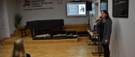 Zdjęcie przedstawia mgr Agatę Bartoś, która prowadzi wykład w auli szkoły. W tle cztery rodzaje saksofonu, fortepian oraz ekran do projekcji. Kolorystyka zdjęcia jest brązowo biała. 