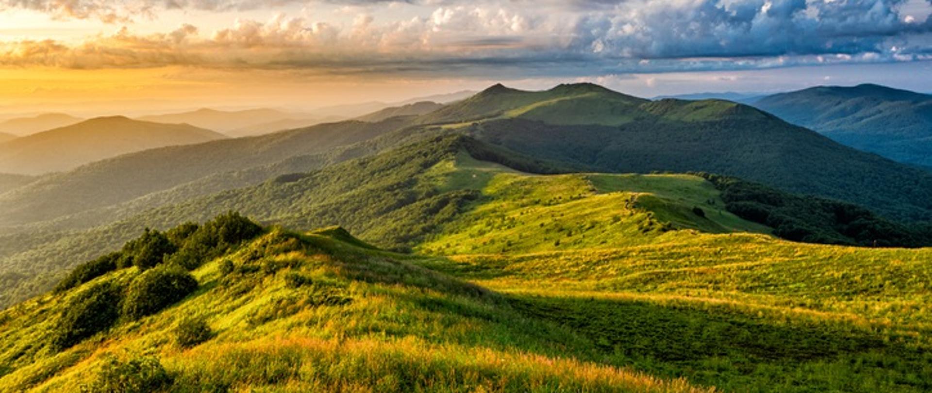 
Szczyty gór Połonina Wetlińska, Bieszczadzki Park Narodowy, Polska. Na górze zdjęcia widoczne chmury, poniżej wierzchołki gór, a najpierwszym planie zielona trawa. Przez chmury przebija się słońce. 