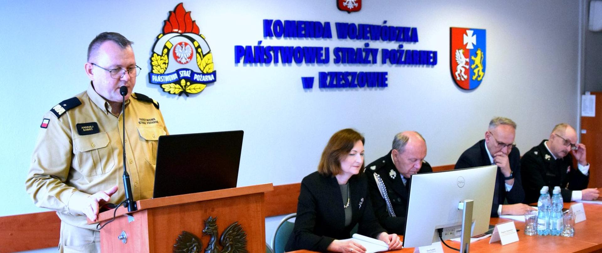 Zdjęcie zrobione w pomieszczeniu, podczas narady podsumowującej działanie strażaków województwa podkarpackiego. Przed mównicą stoi oficer Państwowej Straży Pożarnej w stopniu generała i przemawia. Obok, przy stole siedzą pozostali uczestnicy narady. Na ścianie umieszczony jest logotyp Państwowej Straży Pożarnej herb województwa podkarpackiego oraz napis Komenda Wojewódzka Państwowej Straży Pożarnej w Rzeszowie. 