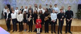Zdjęcie przedstawia grupę uczniów, którzy wzięli udział w koncercie “Młodzi Pianiści Akompaniują”. Uczniowie stoją przed sceną sali koncertowej w szkole, trzymają w rękach instumenty. Na ścianie sali koncertowej wiszą fioletowe lampki choinkowe, bombki i gwiazdy.