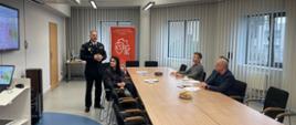 Zdjęcie przedstawia spotkanie w biurze Państwowej Straży Pożarnej. W centrum uwagi znajduje się Komendant Powiatowy PSP w Lublińcu, st. bryg. Janusz Bula.
Przy stole obecni są uczestnicy narady z Rady Powiatu Lublinieckiego. Na ekranie wyświetlana jest prezentacja, w tle baner z logo Krajowego Systemu Ratowniczo – Gaśniczego.