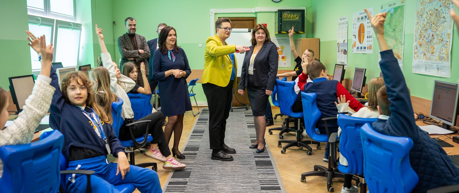 Widok z przodu na klasę, w której pod ścianą i oknami ustawione są stanowiska komputerowe przy których siedzą uczniowie. Część z nich ma podniesioną rękę do góry. Na środku klasy stoi wiceminister Lubnauer z dwoma kobietami i rozmawia.