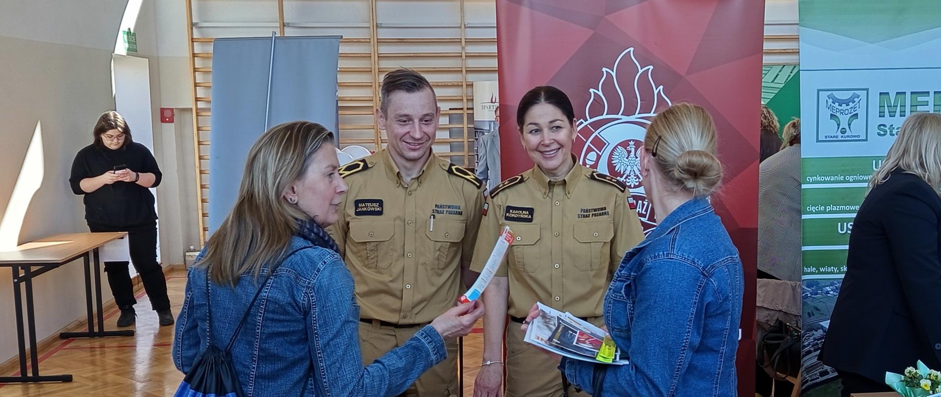 Fotografia przedstawia stoisko Państwowej Straży Pożarnej zorganizowane w sali gimnastycznej podczas targów pracy i edukacji. Na fotografii dwoje funkcjonariuszy rozmawia z odwiedzającymi stoisko kobietami.