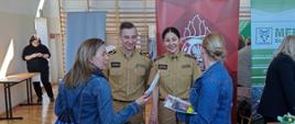 Fotografia przedstawia stoisko Państwowej Straży Pożarnej zorganizowane w sali gimnastycznej podczas targów pracy i edukacji. Na fotografii dwoje funkcjonariuszy rozmawia z odwiedzającymi stoisko kobietami.