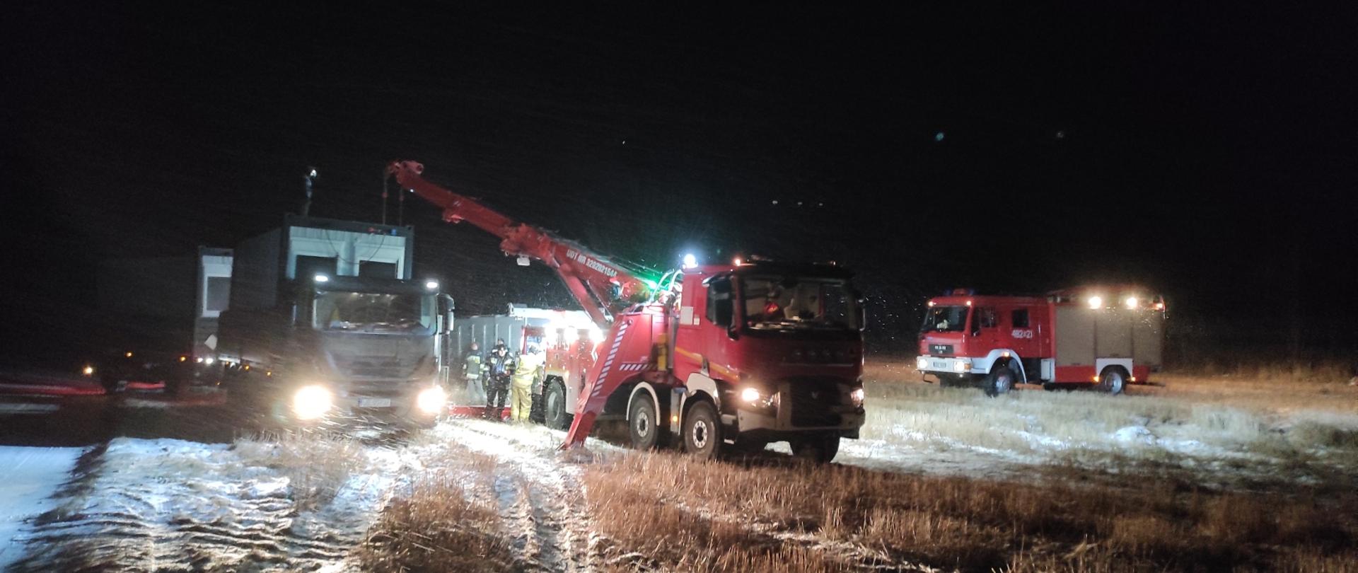Samochód ratownictwa technicznego i strażacy w trakcie rozładunku kontenerów mieszkalnych z przyczepy samochodu wojska polskiego, w tle panuje zmrok