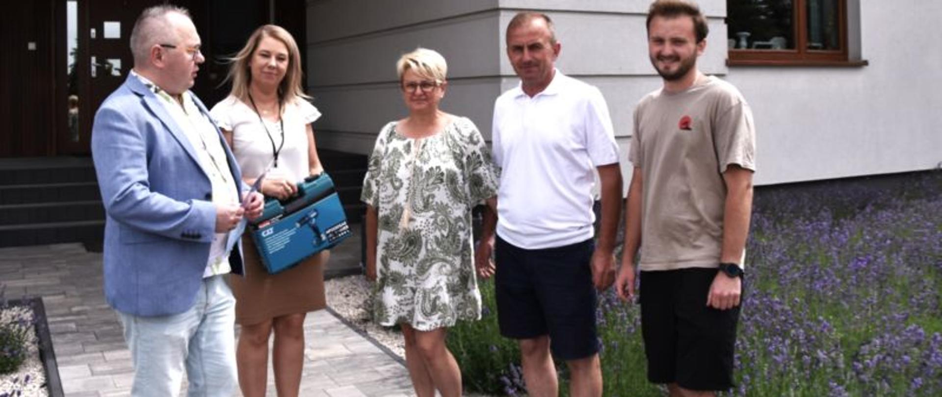  W tle dom a przed nim na chodniku wyłożonym kostką z nasadzeniami lawendy stoją : mężczyzna w marynarce i okularach trzymający w ręku dyplom, po jego lewej stronie kobieta w długich blond włosach w bluzce i spódnicy trzymająca w ręku walizkę z wiertarko-wkrętarką. Po lewej stronie kobiety stoją kolejno laureaci konkursu tj. kobieta w krótkich włosach w sukience, mężczyzna w koszulce polo i krótkich spodenkach oraz ich syn w ubrany w t-shirt i krótkie spodenki