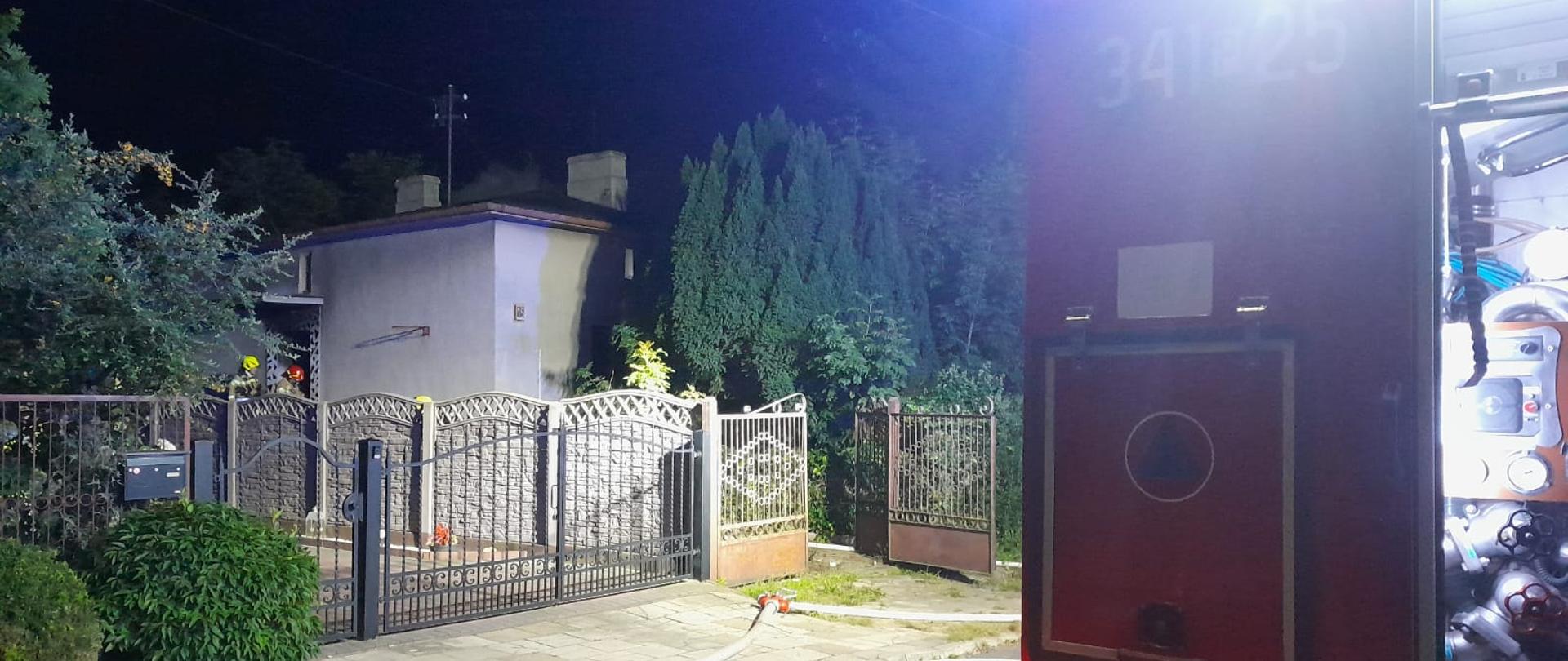 Na zdjęciu wykonanym w porze nocnej widać pojazd straży pożarnej w kolorze czerwonym. Zdjęcie zrobione przed budynkiem w którym powstał pożar. Teren oświetlany jest sztucznym światłem. Widać ulice i po bokach zielone zarośla.