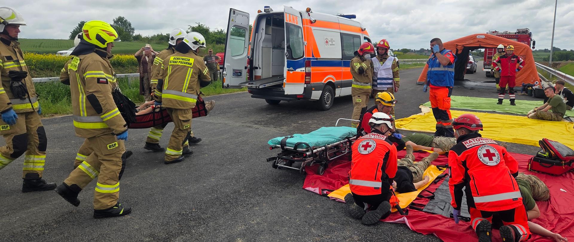 Ćwiczenia ratownicze - na drodze strażacy niosą rannego do ambulansu, obok ratownicy udzielają pierwszej pomocy leżącym na jezdni na czerwonej płachcie 