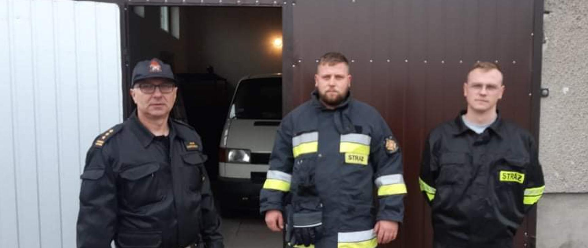 Trzech mężczyzn stoi na tle brązowej bramy garażowej budynku OSP. Przez otwarte drzwi widać kawałek białego samochodu. Mężczyzna po lewej to Komendanta PSP w Żurominie. Dwóch pozostałych to druhowie OSP. 