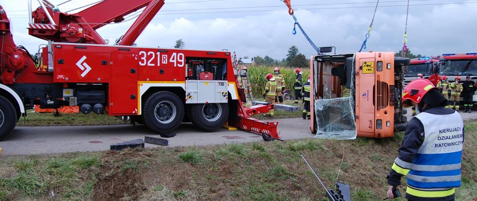Na zdjęciu pokazane stawianie na koła rozbitego w wypadku autokaru. Działania prowadzone z wykorzystaniem ciężkiego samochodu ratownictwa technicznego. Na pierwszym planie, po lewej stronie strażak w ubraniu specjalnym oraz w kamizelce z napisem kierujący działaniem ratowniczym.