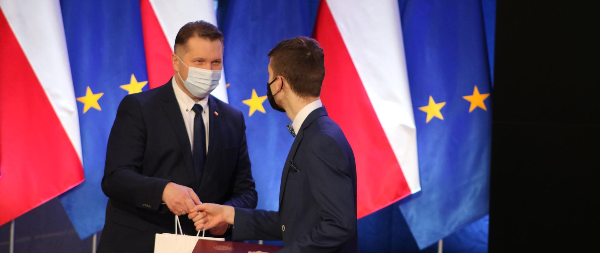 Minister Przemysław Czarnek wręcza upominek i dyplom chłopcu ubranemu w garnitur. Obaj mają na twarzach maseczki. W tle flagi polskie i unijne. 