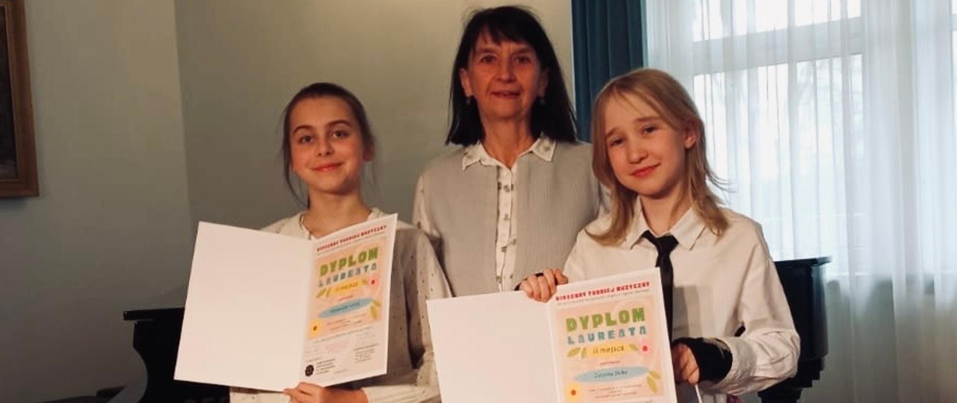 Na zdjęciu widać dwie dziewczynki stojące obok nauczycielki w eleganckim, jasnym wnętrzu z fortepianem w tle. Dziewczynki trzymają w rękach dyplomy oraz torby z upominkami, a na ich twarzach widać dumę i radość. Nauczycielka stojąca pośrodku uśmiecha się z satysfakcją. Cała trójka ubrana jest schludnie, w jasnych koszulach, co nadaje zdjęciu uroczysty charakter.
