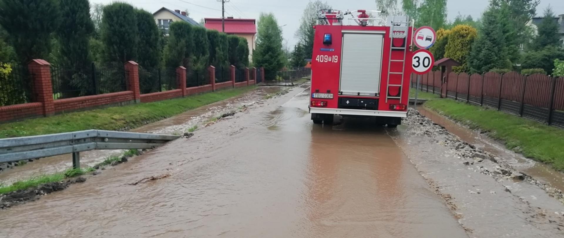 Na zdjęciu woda przepływająca przez drogę asfaltową, w tle pojazd pożarniczy