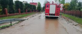 Na zdjęciu woda przepływająca przez drogę asfaltową, w tle pojazd pożarniczy