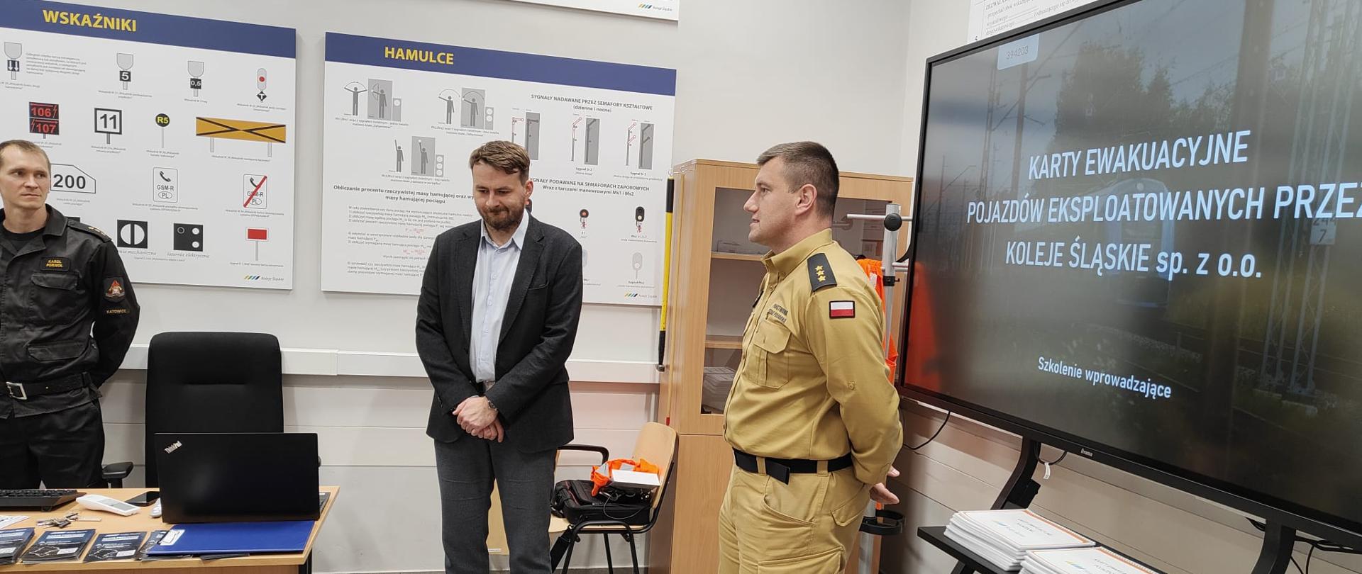 Na zdjęciu sala wykładowa. Na ścianie wiszą tablice informacyjne. Po prawej stronie duży monitor telewizyjny, na którym wyświetla się napis "Karty ewkuacyjne pojazdów eksploatowanych przez Koleje Śląskie sp. z o.o." Po lewej stronie stoi biurko, na nim laptop i ulotki. Na środku sali, tyłem do telewizora stoi strażak w piaskowym mundurze służbowym, obok niego stoi meżczyzna w marynarce. Za biurkiem stoi strażak w czarnym mundurze służbowym.