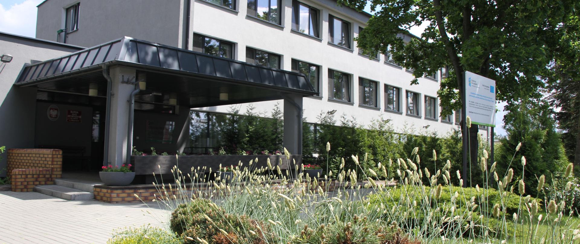 Zdjęcie przedstawia jasnoszary budynek bursy od strony ul. Muzycznej. Pośrodku wejście główne do budynku z ciemnoszarym zadaszeniem wpartym na dwóch betonowych słupach i sektor A po prawej stronie. Przy wejściu donice z kwiatami, murek i dwa schodki. Ponad to po prawej stronie drzewo i tablica informacyjna. z przodu kwitnące trawy i krzewy.