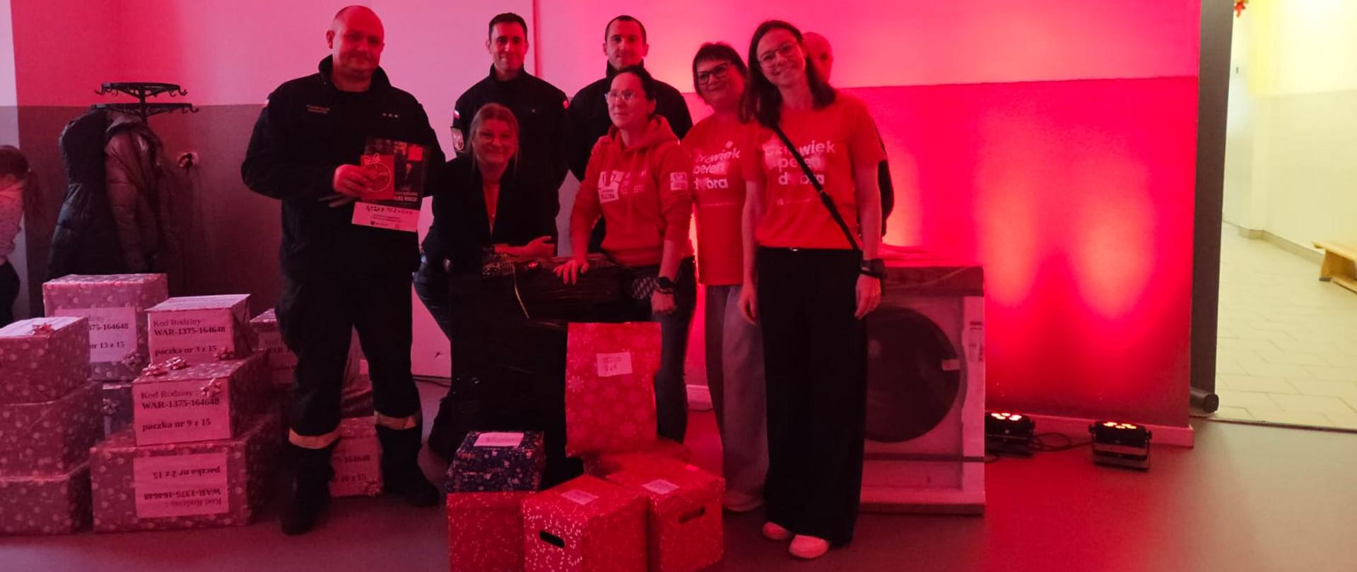 Zdjęcie przedstawia wolontariuszy akcji „Szlachetna Paczka” oraz funkcjonariuszy Komendy Powiatowej Państwowej Straży Pożarnej w Gołdapi obok upominków przekazanych przez strażaków, zlokalizowanych w kwaterze głównej akcji „Szlachetna Paczka” w powiecie gołdapskim. Obraz uchwycony w innej konfiguracji niż poprzednie zdjęcie