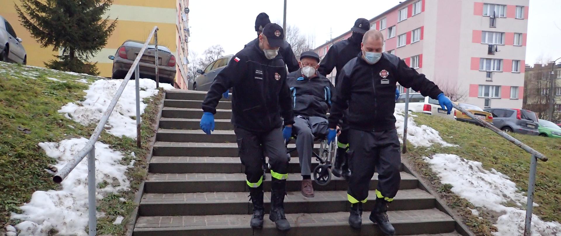 Czterech druhów OSP znoszących mężczyznę na wózku inwalidzkim ze schodów na osiedlu mieszkalnym.