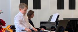 Chłopczyk gra pałkami na ksylofonie, w tle kobieta gra na fortepianie.