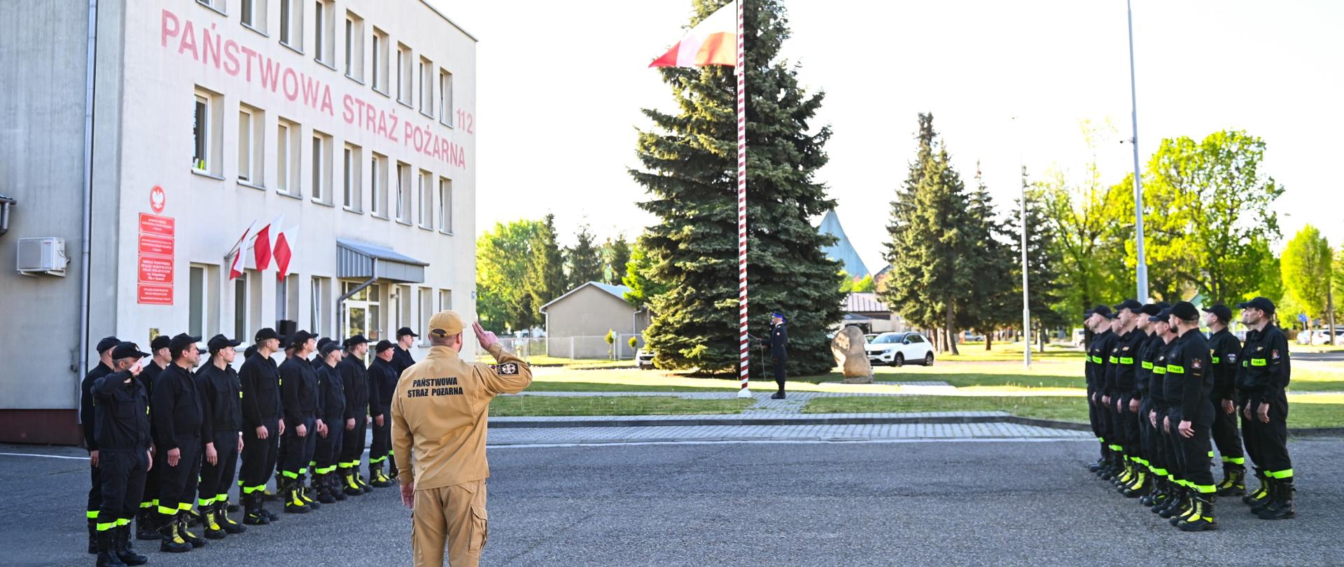 Na placu przed Jednostka Ratowniczo Gaśnicza nr 1 stoją strażacy w dwóch szpalerach naprzeciw siebie każdy szpaler w dwuszeregu na uroczystej zbiórce, ubrani w czarne ubrania koszarowe. Na środku stoi dowódca ubrany w piaskowy mundur. Na maszt uroczyście wciągana jest biało czerwona flaga przez poczet sztandarowy ubrany w mundury galowe.