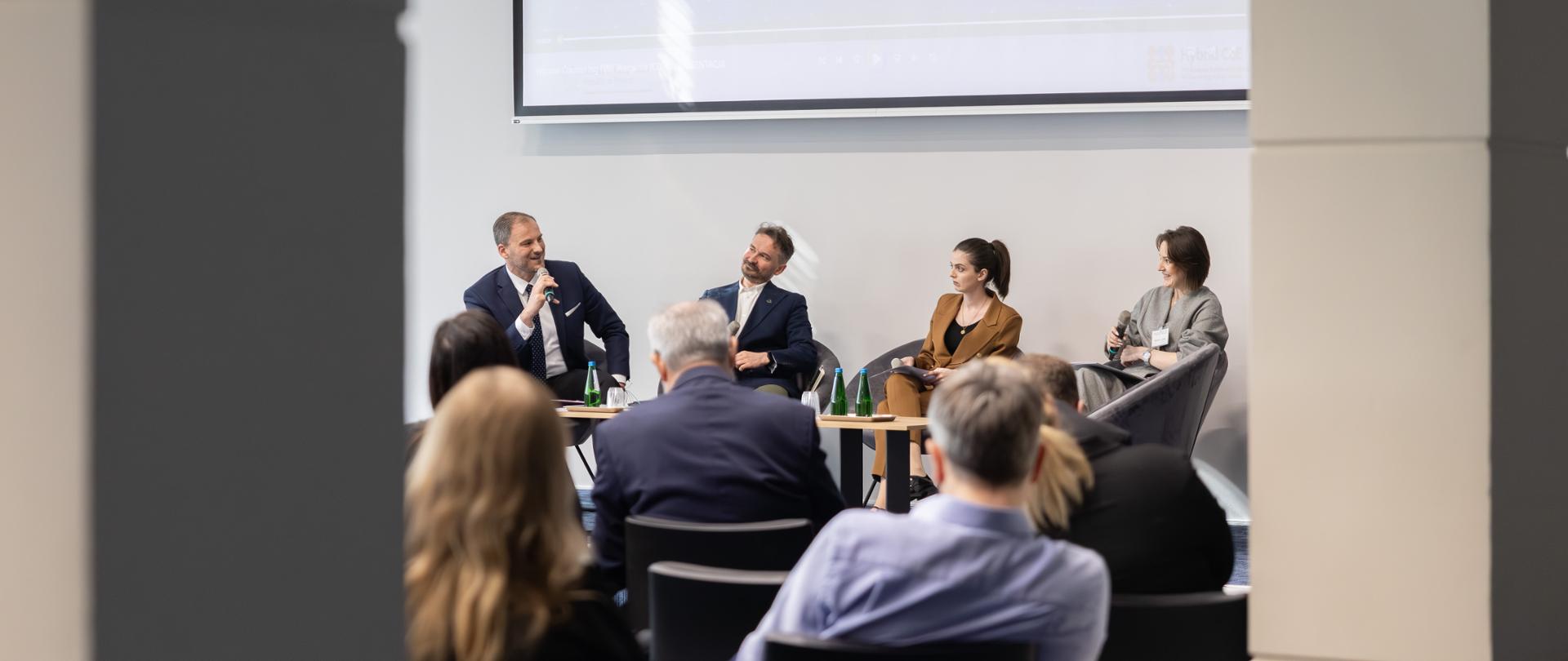 Panel discussion at a conference – two men and two women in formal attire sit on stage and speak, one of the men holding a microphone; audience members are seated in front.