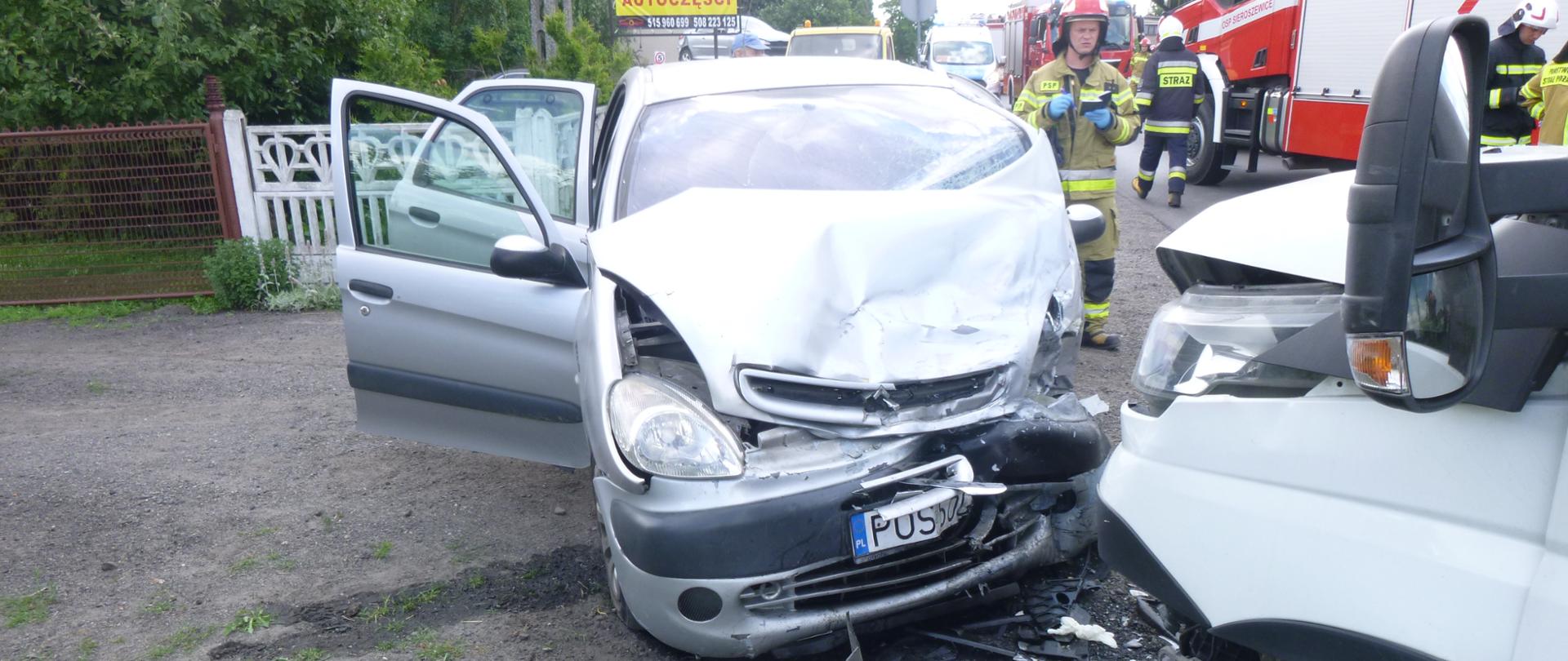 Na pierwszym planie widać zderzenie dwóch pojazdów osobowych. W tle przygląda im się jeden strażak, pozostali strażacy znajdują się w pobliżu oddalonych wozów strażackich.