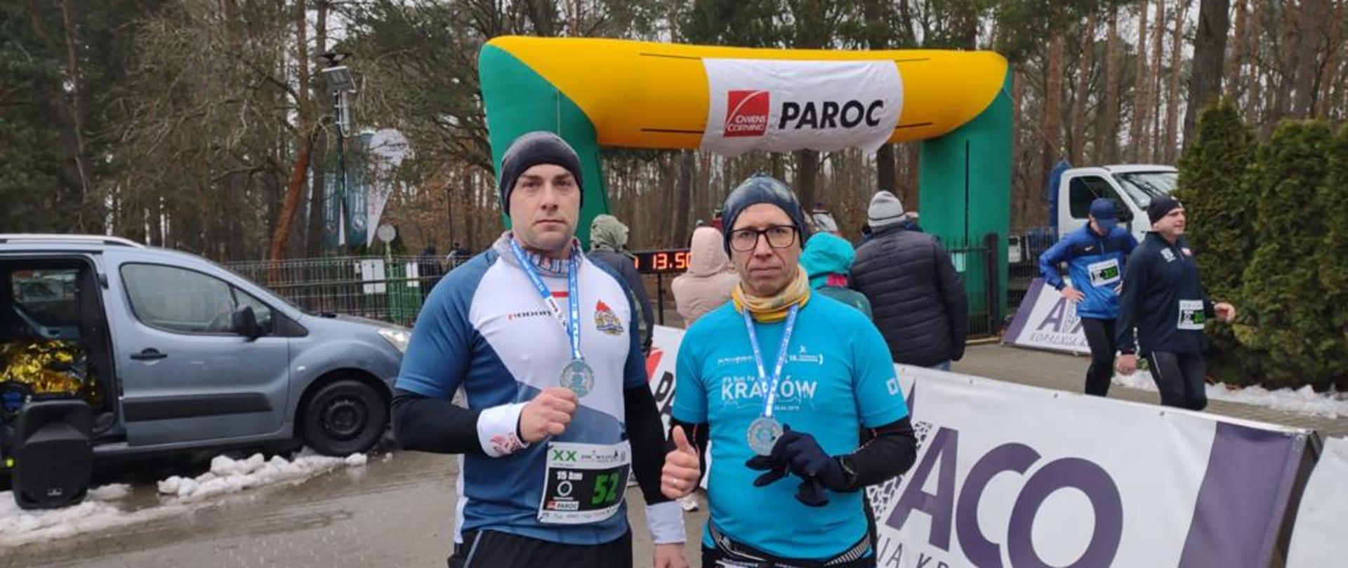 Dwóch mężczyzn w sportowych strojach stoi przed bramką mety, każdy z nich pokazuje zdobyty medal