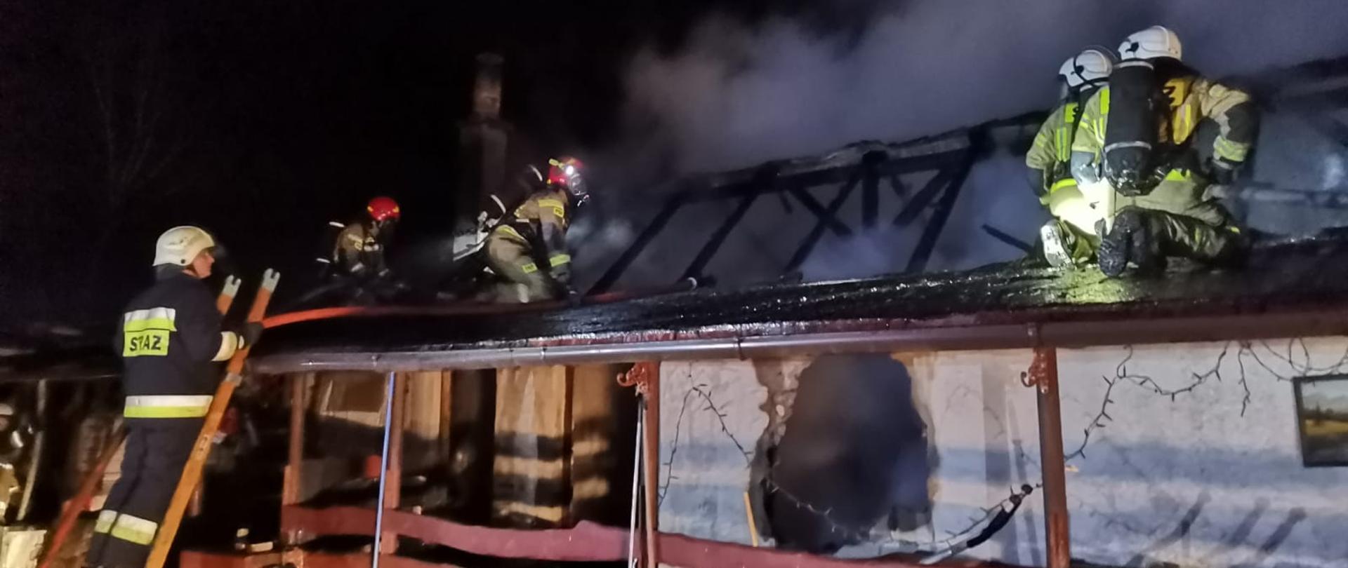 Fotografia przedstawia ujęcie na wprost budynku jednokondygnacyjnego z częściowo spalonym poszyciem dachu. Widoczne są zwęglone krokwie. Na przedniej ścianie widoczna duża dziura średnicy około 1 metra. Na dachu nad krużgankiem widać dwie roty strażaków z liniami gaśniczymi, ubranych w ubrania specjalne, w hełmach, z założonymi aparatami powietrznymi. O krużganek oparta jest drabina nasadkowa na której stoi strażak w ubraniu specjalnym i w hełmie. Jest ciemno, budynek jest oświetlony z zewnątrz przez najaśnice.