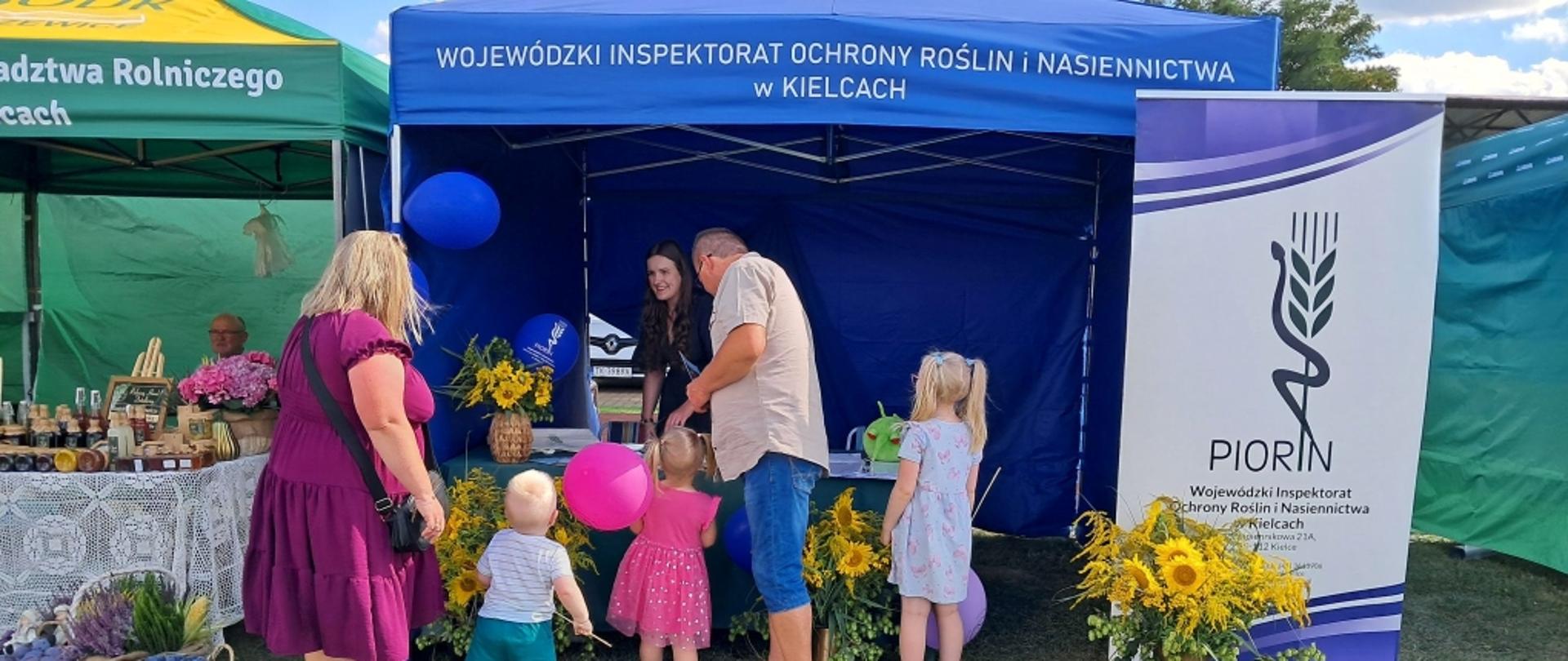 Rodzina z trójką małych dzieci odwiedza niebieskie stoisko Wojewódzkiego Inspektoratu Ochrony Roślin i Nasiennictwa w Kielcach ozdobione bukietami słoneczników.