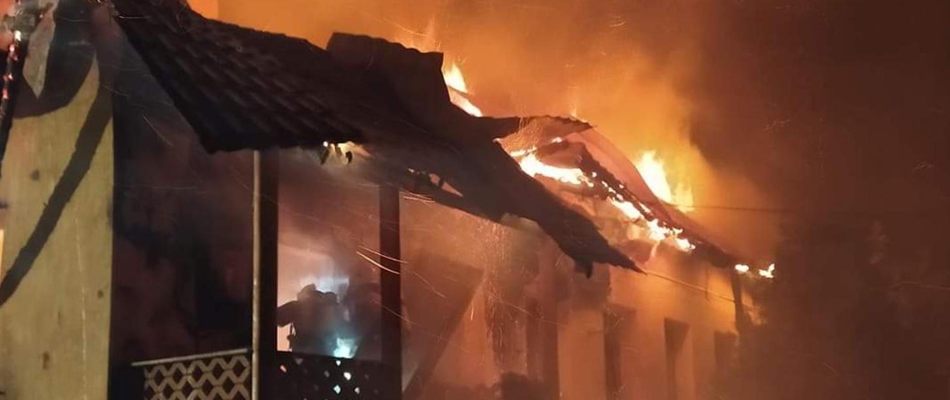 Zdjęcie nocne przedstawiające pożar parterowego budynku mieszkalnego jednorodzinnego. Płomienie wydobywają się z pod pokrycia dachu. W wejściu do płonącego budynku widoczny jest strażak w ubraniu specjalnym, wyposażony w aparat powietrzny. 