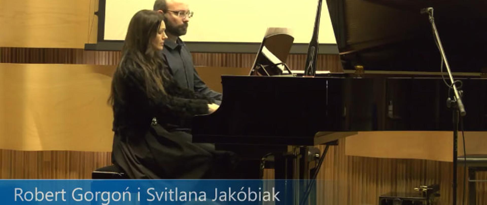 Na tle paneli akustycznych oraz ekranu do projektora dwóch pianistów gra na cztery ręce przy otwartym fortepianie.