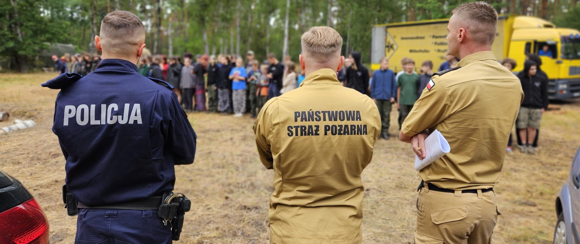 Trzech funkcjonariuszy – dwóch z Państwowej Straży Pożarnej i jeden z Policji – stoi tyłem, obserwując grupę młodzieży zgromadzoną na leśnej polanie. W tle widoczna ciężarówka i las.