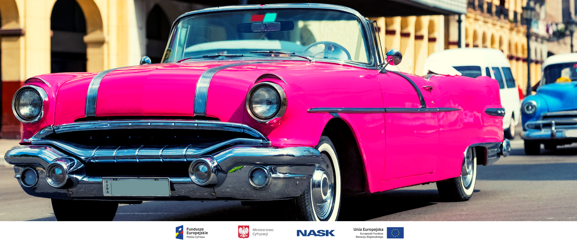 Na pierwszym planie zabytkowy różowy kabriolet stojący na ulicy. W tle budynek z arkadami i inne zabytkowe samochody