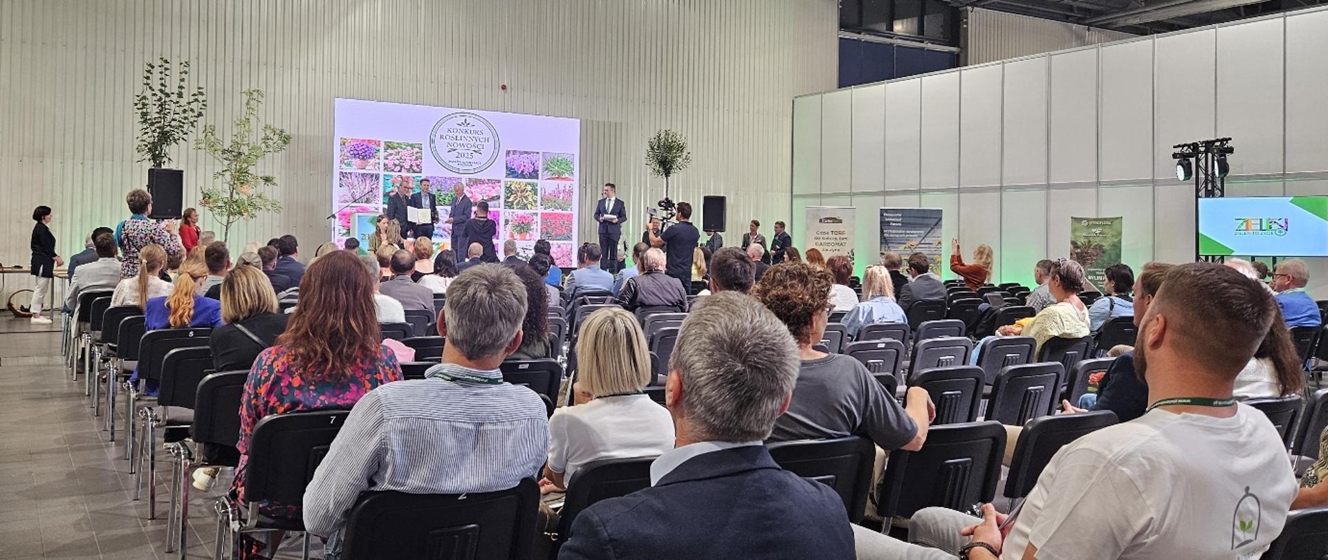 Publiczność siedzi na sali podczas konferencji branżowej, na scenie odbywa się prezentacja z udziałem prelegentów; w tle widoczny napis „KONKURS ROŚLINNYCH NOWOŚCI 2025”.