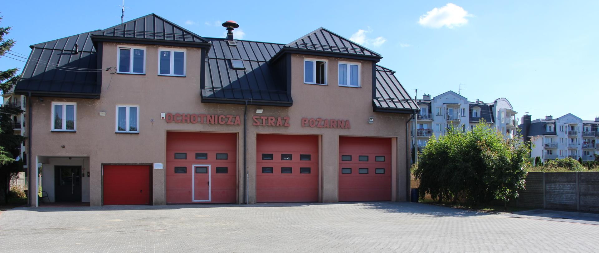 Strażnica Ochotniczej Straży Pożarnej w Jabłonnie. NA zdjęciu budynek piętrowy. Na parterze 4 bramy na pojazdy bojowenad bramami napis ochotnicza straż pożarna dach szary.