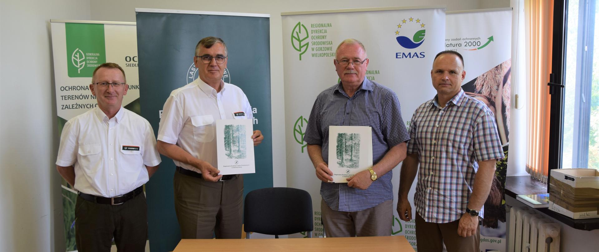 Czterech mężczyzn pozuje do zdjęcia. Dwóch stojących w środku trzyma w rękach białe teczki z rysunkiem lasu. Przed nimi stoi stół, na nim leżą dwa długopisy.