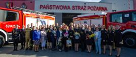 Strażacy w mundurach i osoby cywilne stoją pozując do zdjęcia pomiędzy dwoma ustaionymi w literę V wozami strażackimi. Nad ich głowami napis Państwowa Straż Pożarna.