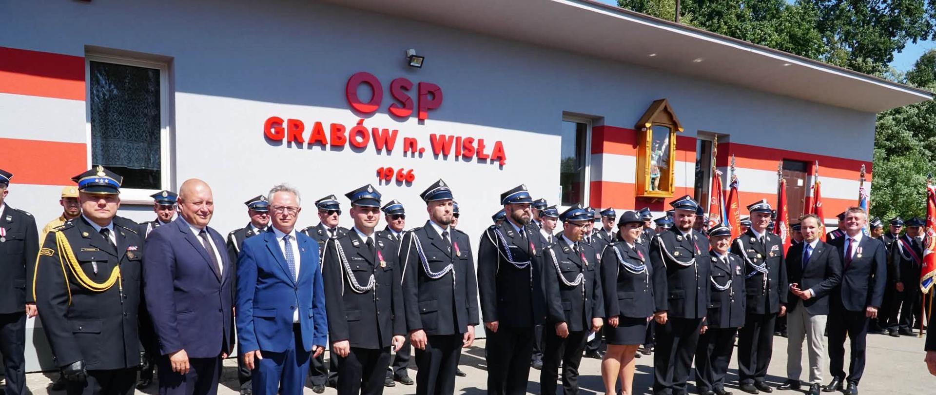 Fotografia grupowa wykonana przed budynkiem Ochotniczej Straży Pożarnej w Grabowie nad Wisłą. W pierwszym rzędzie stoi trzech mężczyzn w garniturach oraz liczna grupa strażaków w galowych, ciemnogranatowych mundurach z czapkami rogatywkami. Za nimi, w tle, widoczny jest jasnoszary budynek remizy z czerwonymi pasami. Nad wejściem widnieje duży, czerwony napis: "OSP GRABÓW n. WISŁĄ 1966". Po prawej stronie, na ścianie, wisi kapliczka z figurą św. Floriana. Strażacy w dalszych rzędach trzymają sztandary. Dzień jest słoneczny, niebo bezchmurne.