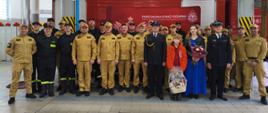 Zdjęcie grupowe w garażu JRG przedstawia uczestników uroczystej zbiórki. Strażacy w ubraniach koszarowych, służbowych oraz zastępca komendanta i odchodzący na emeryturę funkcjonariusz w mundurach.