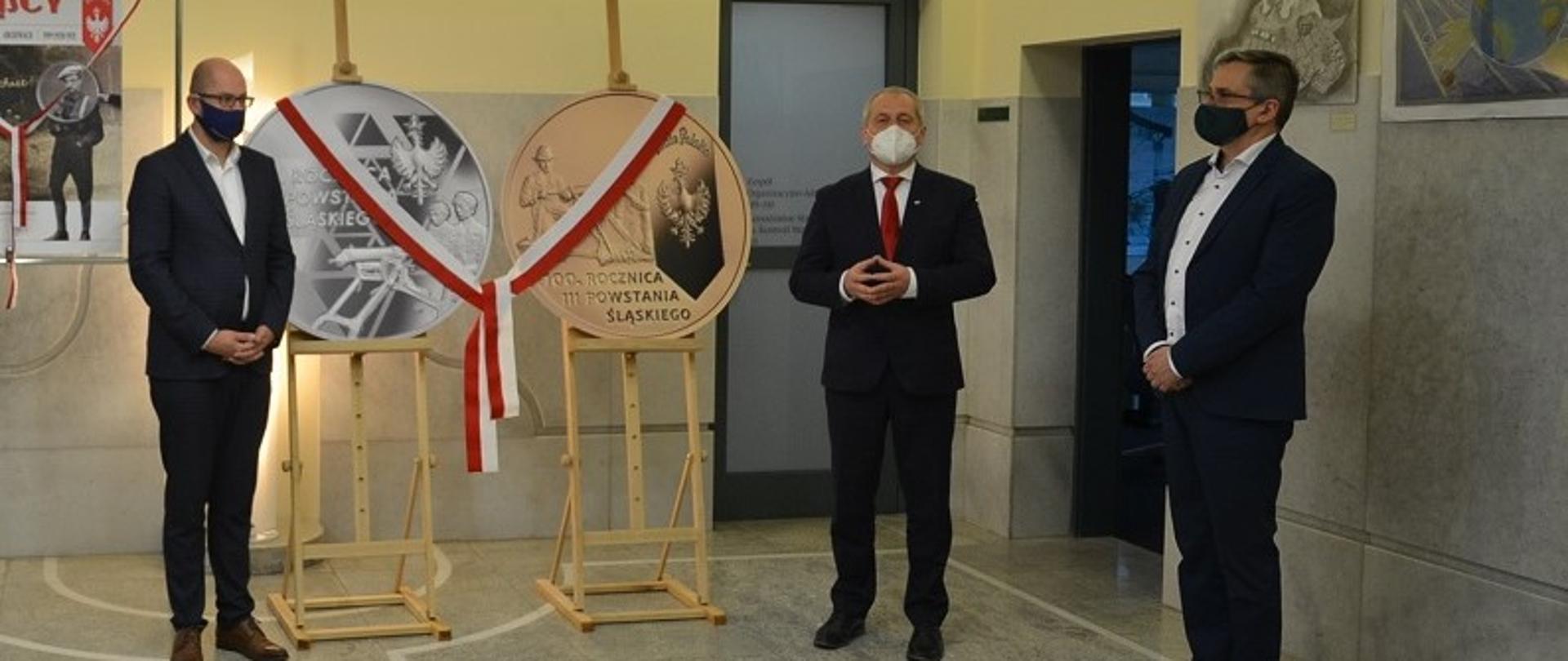 Na zdjęciu trzech mężczyzn. Od lewej wicemarszałek woj. opolskiego, wojewoda opolski i dyrektor opolskiego oddziału NBP. Mężczyźni stoją od siebie w dystansie, za ich plecami na drewnianym stelażu znajdują się podobizny dwóch moment: srebrnej i złotej. Monety te przepasane są biało-czerwoną wstęgą. W tle granitowa ściana. 