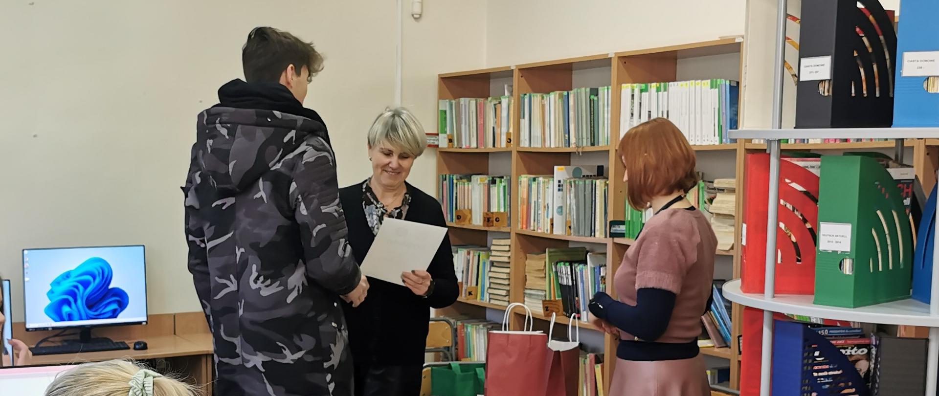 W sali biblioteki szkolnej stoją trzy osoby, kobieta wręcza dyplom uczniowi.

