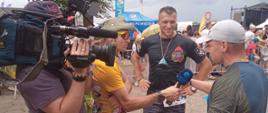 Na zdjęciu widać uczestnika biegu który udziela wywiadu przed kamerą. Na drugim planie widać operatora kamery oraz prowadzącego wywiad w tle stoi drugi uczestnik. 