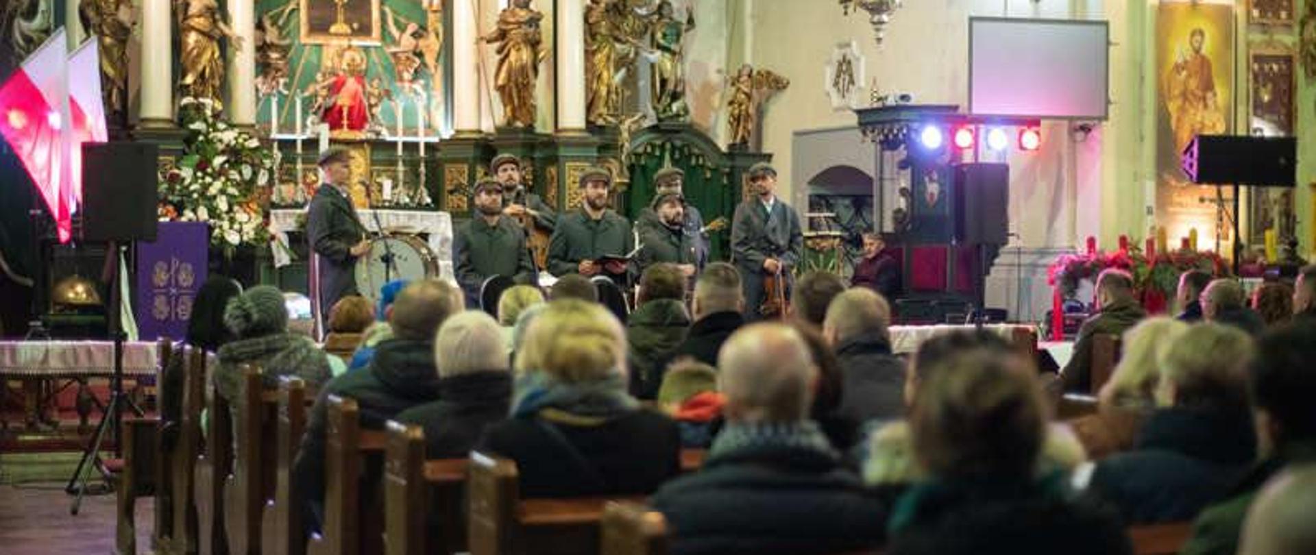 Wnętrze zabytkowego kościoła. Na tle ołtarza stoi zespół złożony z siedmiu osób, przebrani w stare munduru wojskowe. W ławkach siedzą osoby które słuchają koncertu. Z lewej strony wiszą dwie Polskie flagi.
